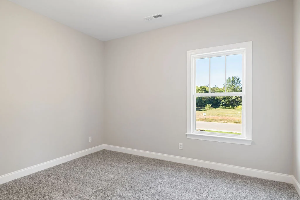 Empty room with light gray walls, carpeted floor, and a window showing outdoor greenery. Design by Pratt Home Builders in Chattanooga TN.