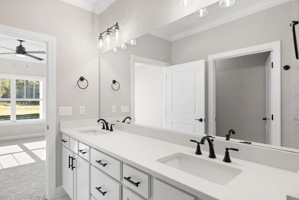 Modern bathroom with dual sinks, black fixtures, large mirror, and white cabinets. Sunlit bedroom view. Pratt Home Builders in Chattanooga TN.
