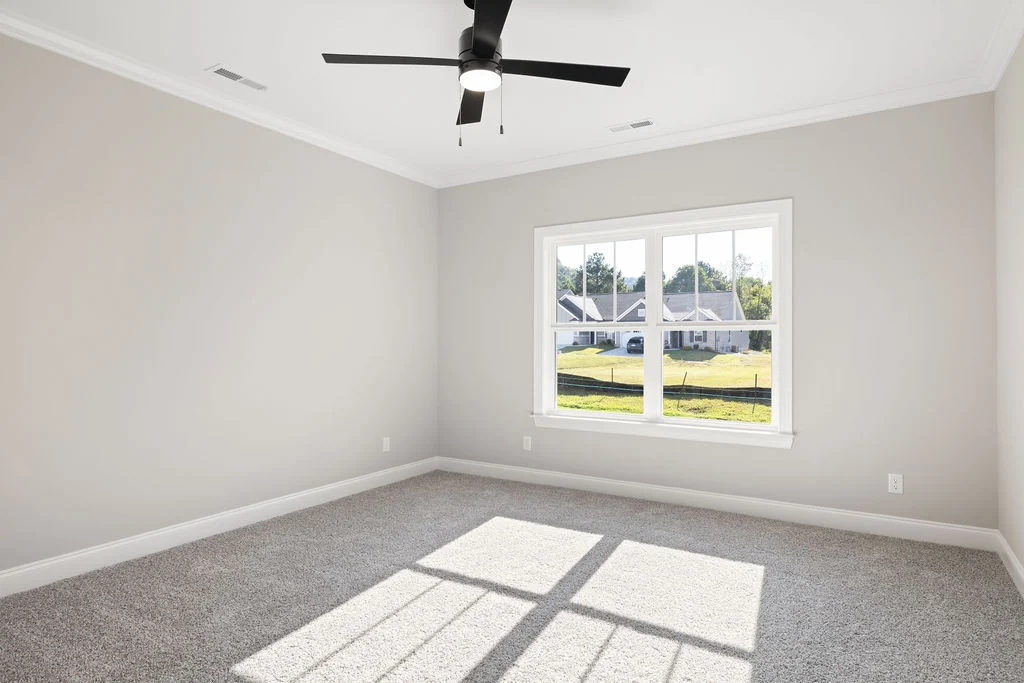 Empty, sunlit room with beige walls, large window, ceiling fan, and gray carpet. Built by Pratt Home Builders in Chattanooga, TN.