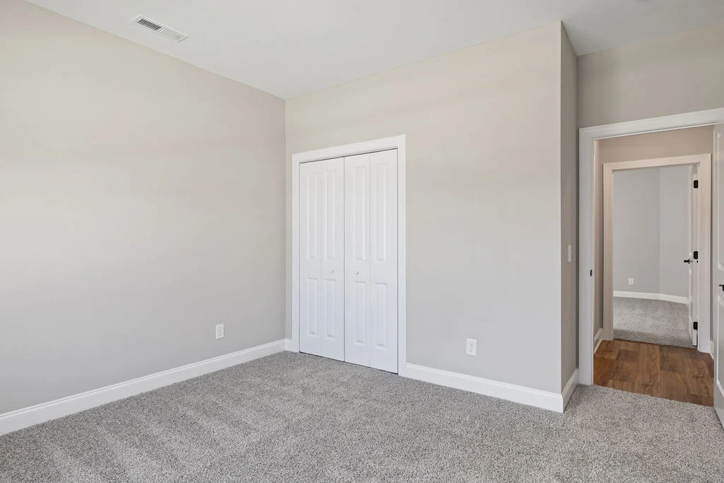 Empty room with gray carpet, beige walls, and white closet doors. Adjacent hallway and hardwood floor. Built by Pratt Home Builders in Chattanooga TN.