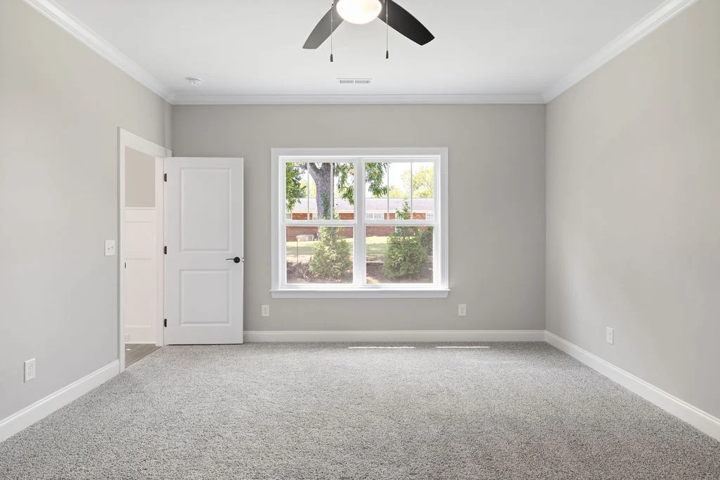 Bright, empty room with gray carpet, white door, and large window by Pratt Home Builders in Chattanooga TN, featuring neutral wall tones and ceiling f