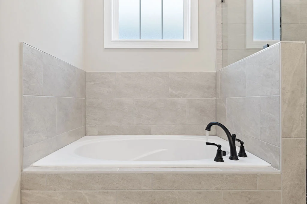 Modern bathtub with elegant black fixtures, surrounded by light gray tiles, under a frosted window. Designed by Pratt Home Builders in Chattanooga TN.