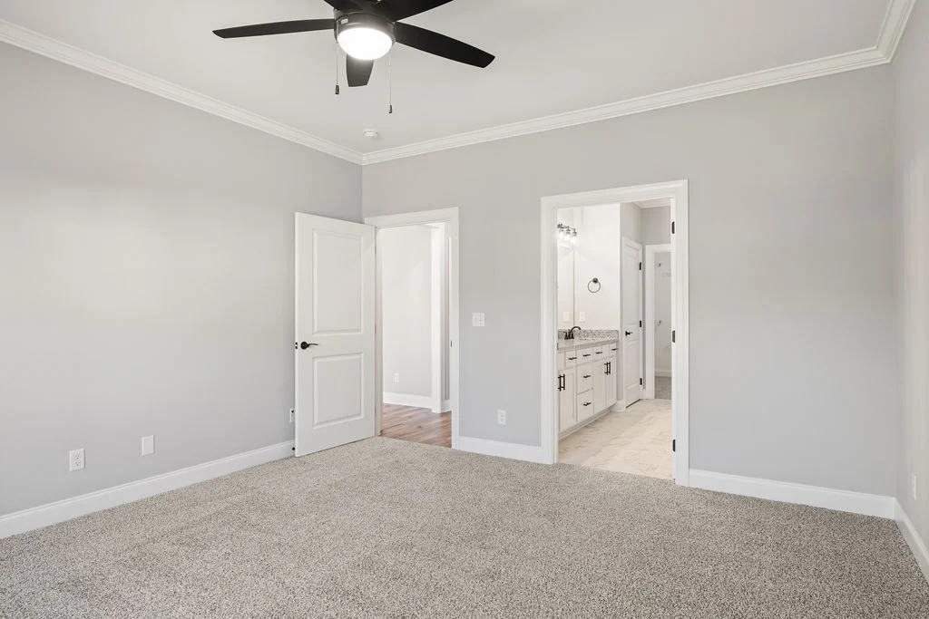 Spacious bedroom with gray walls, carpeted floor, ceiling fan, open doors to hallway and bathroom. Crafted by Pratt Home Builders in Chattanooga TN.