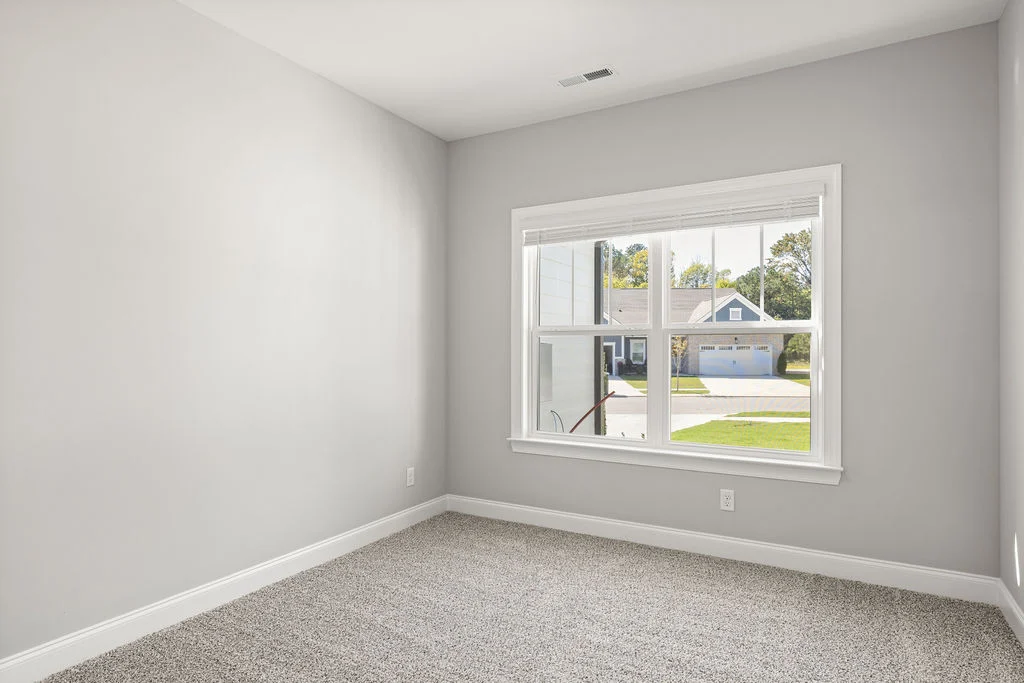 Bright empty room with gray walls, carpeted floor, large window showing suburban street view. Built by Pratt Home Builders in Chattanooga TN.