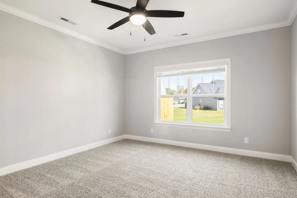 Bright, empty room with gray carpet, large window, and ceiling fan. Modern design by Pratt Home Builders in Chattanooga TN.
