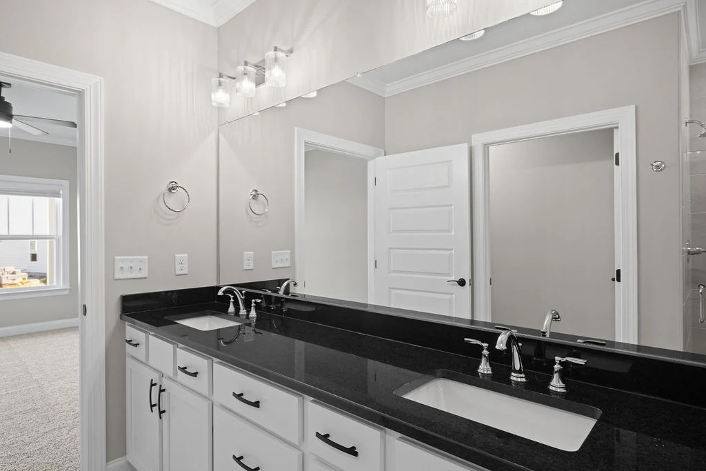 Modern bathroom with double sinks, black countertops, and white cabinets by Pratt Home Builders in Chattanooga TN. Open doorway to carpeted room.