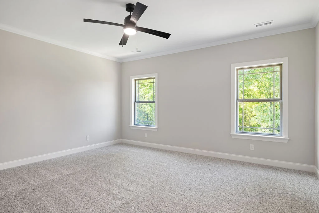 Spacious, carpeted room with neutral walls, two windows overlooking greenery, and ceiling fan. Built by Pratt Home Builders in Chattanooga TN.