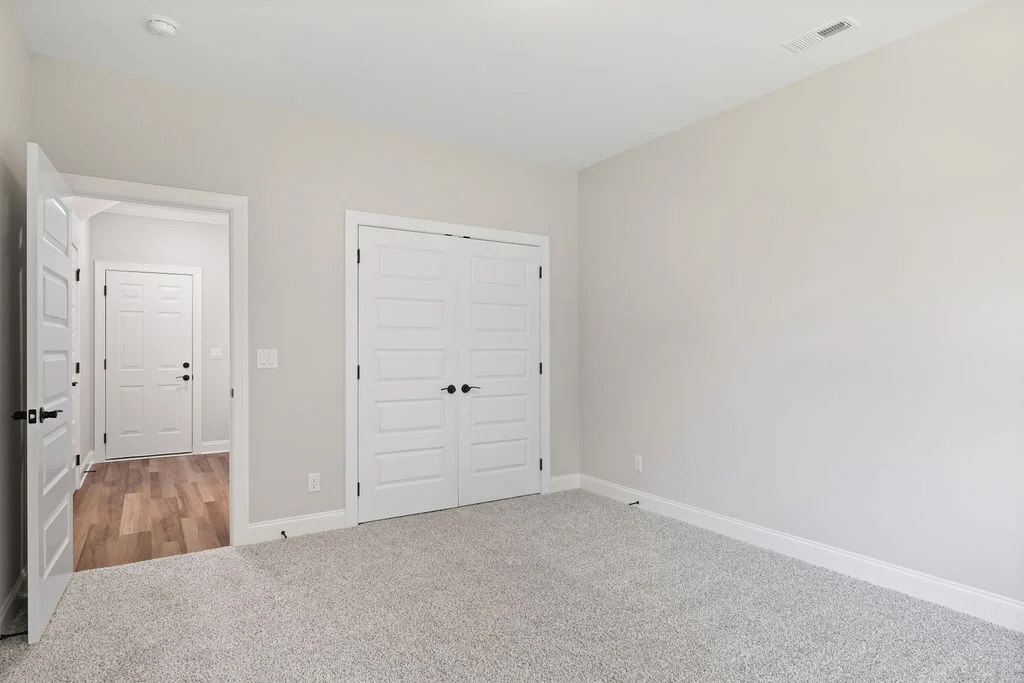 Empty room with neutral walls, textured carpet, and double closet doors. Open door reveals hallway with wood flooring. Built by Pratt Home Builders in