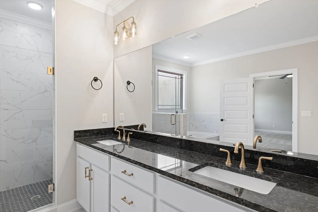 Modern bathroom by Pratt Home Builders in Chattanooga TN, features a double vanity with black countertops, gold fixtures, and a glass shower door.