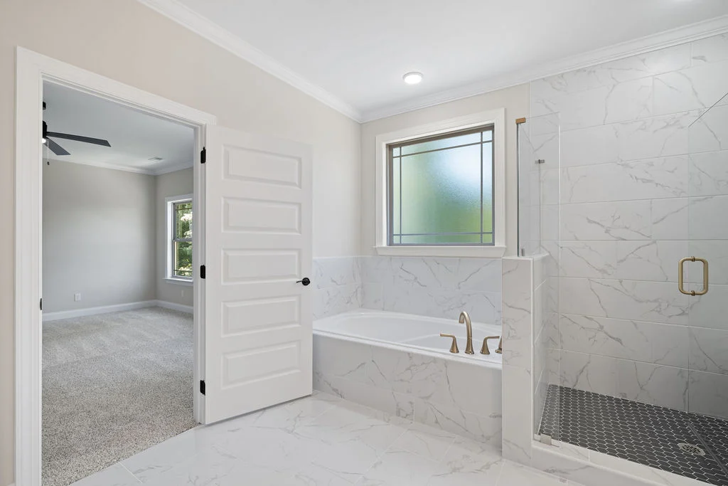 Spacious bathroom with marble tiles, glass shower, bathtub, and adjacent carpeted room. Pratt Home Builders in Chattanooga TN.
