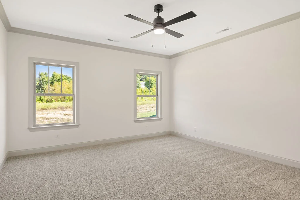 Spacious empty room with carpet, two large windows, ceiling fan, and view of green landscape, designed by Pratt Home Builders in Chattanooga TN.