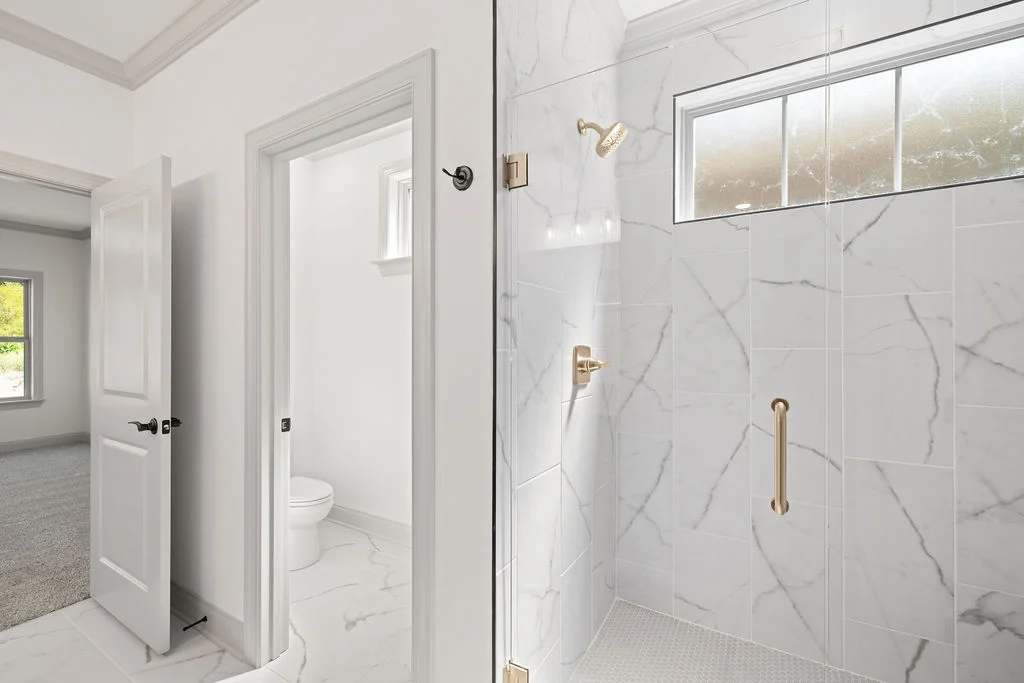 Modern bathroom with marble shower, glass panel, and adjacent open door to toilet area. Designed by Pratt Home Builders in Chattanooga TN.