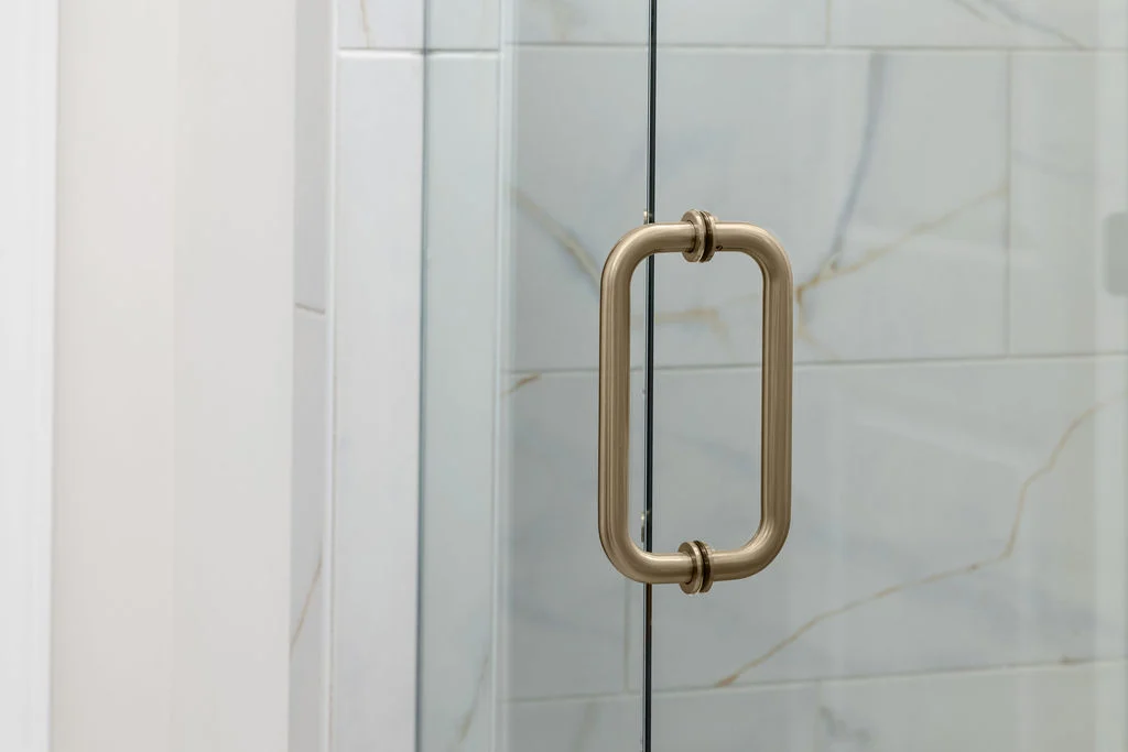 Brushed metal shower door handle on a glass door, surrounded by marble tiles; design by Pratt Home Builders in Chattanooga TN.