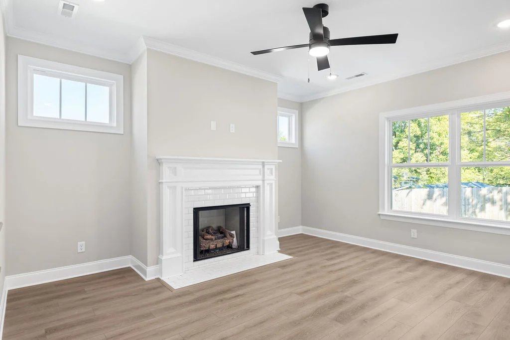 Living room by Pratt Home Builders in Chattanooga TN, featuring a white fireplace, ceiling fan, and large windows with wood flooring.