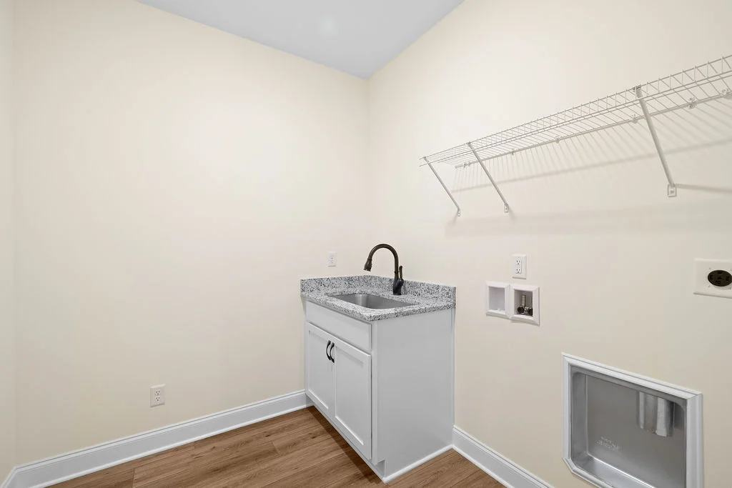Laundry room by Pratt Home Builders in Chattanooga TN, featuring a granite countertop sink, white cabinets, and wire shelf on beige walls.