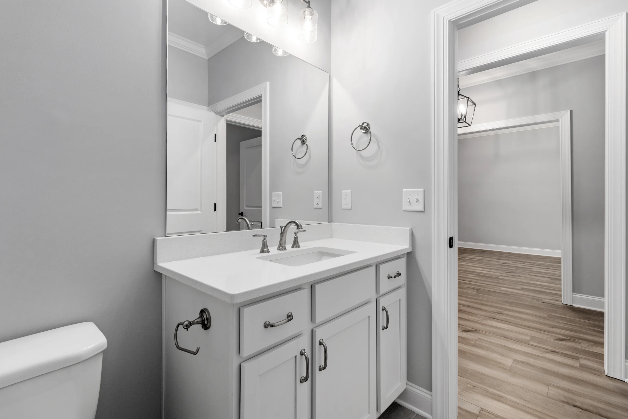 Modern bathroom with white vanity, single sink, and elegant lighting by Pratt Home Builders in Chattanooga TN. Hallway with wooden floor visible.