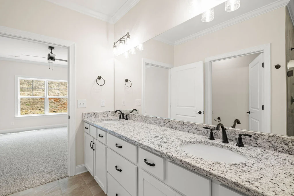Modern bathroom with granite countertop, double sink, and white cabinetry. View into carpeted bedroom. Built by Pratt Home Builders in Chattanooga TN.