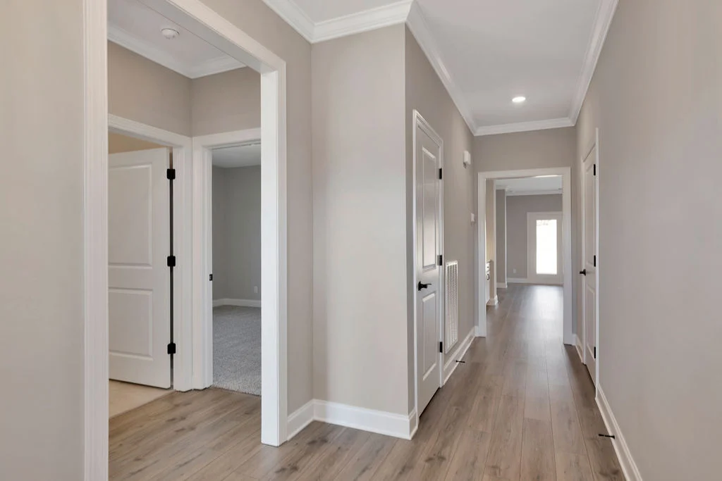 Modern hallway with wood flooring, beige walls, and white trim. View of multiple doors and natural light from the end. Designed by Pratt Home Builders