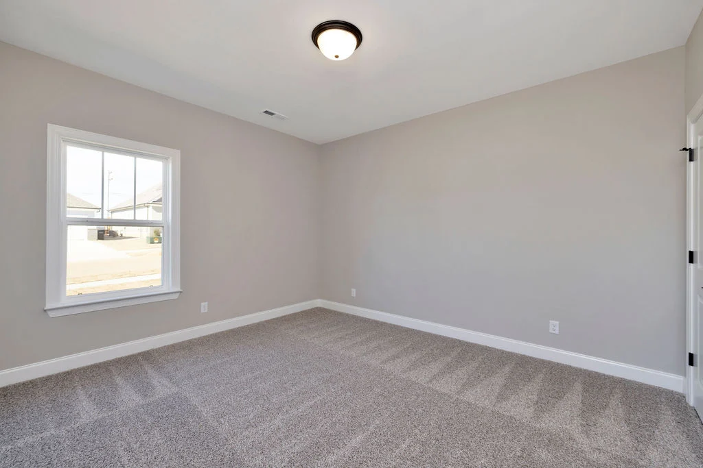 Empty, carpeted room with light gray walls and ceiling light, built by Pratt Home Builders in Chattanooga, TN. Single window on the left wall.