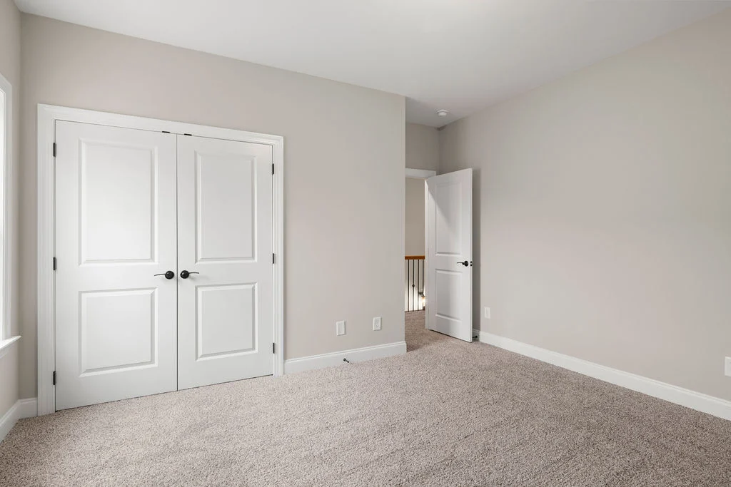 Empty beige carpeted room with closed double closet doors and open entry door, built by Pratt Home Builders in Chattanooga TN.