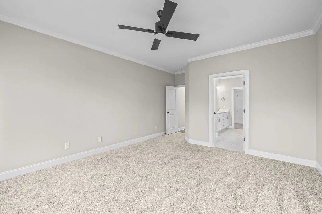 Minimalist bedroom with beige carpet, gray walls, ceiling fan, and open door leading to a bright bathroom. Pratt Home Builders in Chattanooga TN.