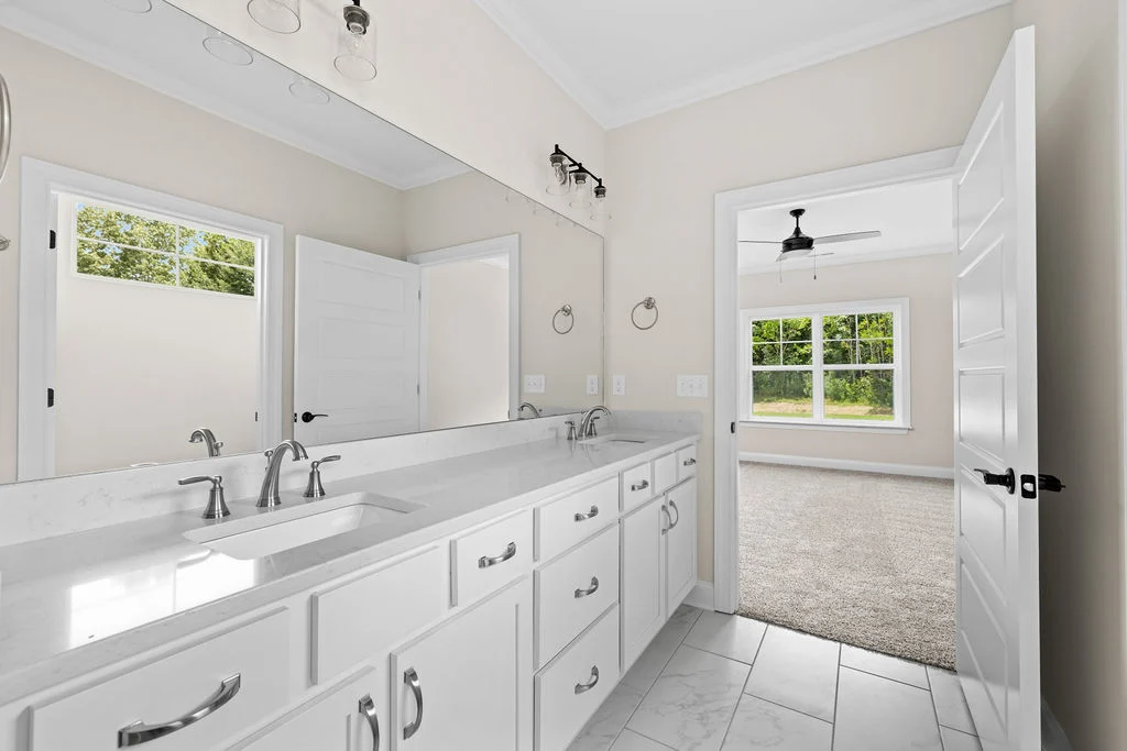 Modern bathroom with dual sinks and large mirror, leading to a bright bedroom with carpet. Designed by Pratt Home Builders in Chattanooga TN.