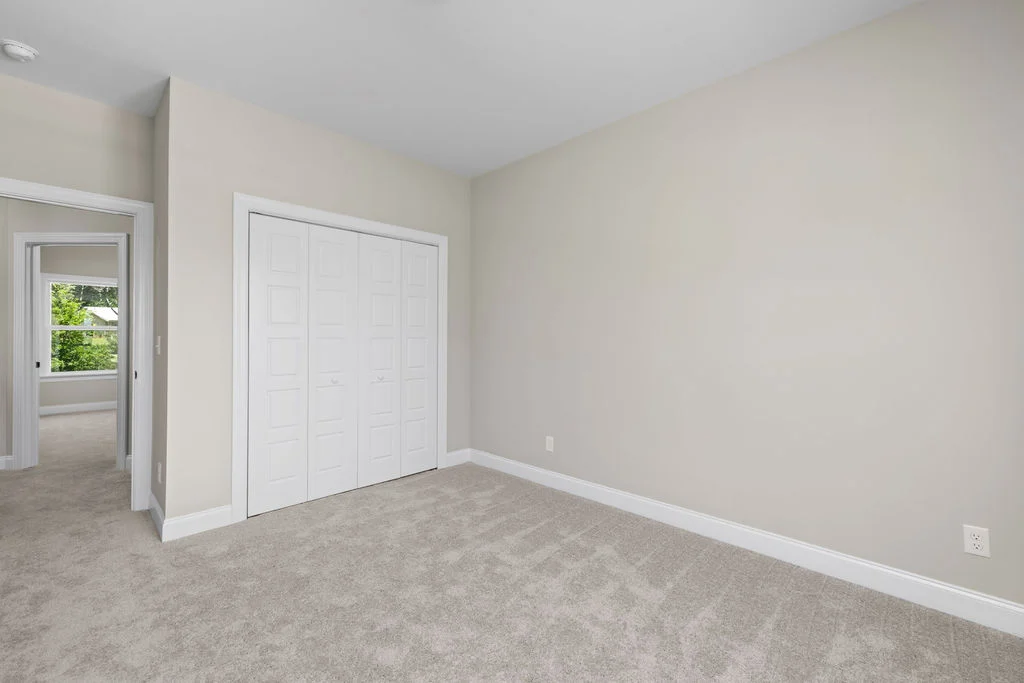 Empty room with beige walls and carpet, white closet doors. Sunlit hallway and window view. Home by Pratt Home Builders in Chattanooga TN.