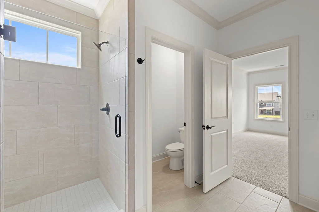 Modern bathroom and bedroom with beige tiles, glass shower, and plush carpet, designed by Pratt Home Builders in Chattanooga TN.