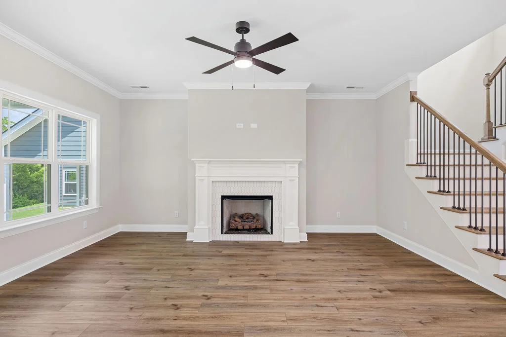 Bright living room with wood floors, white fireplace, large window, ceiling fan, and staircase by Pratt Home Builders in Chattanooga TN.