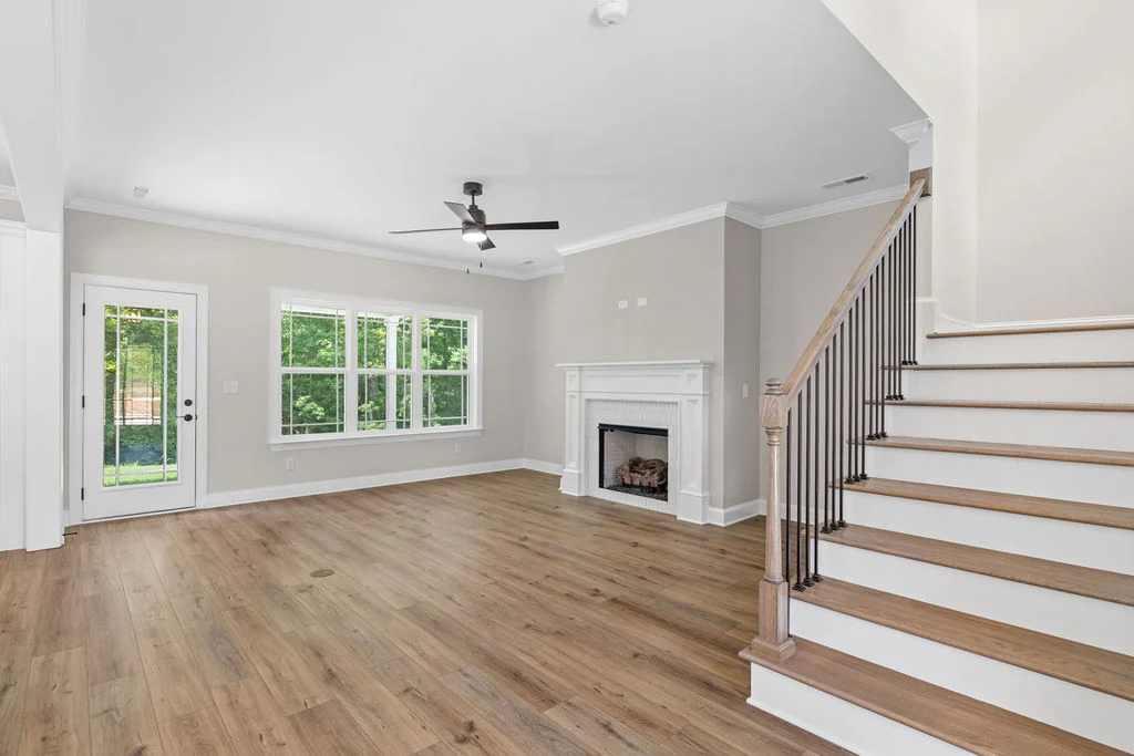 Bright, spacious living room by Pratt Home Builders in Chattanooga TN, featuring wood floors, a fireplace, large windows, and adjacent staircase.