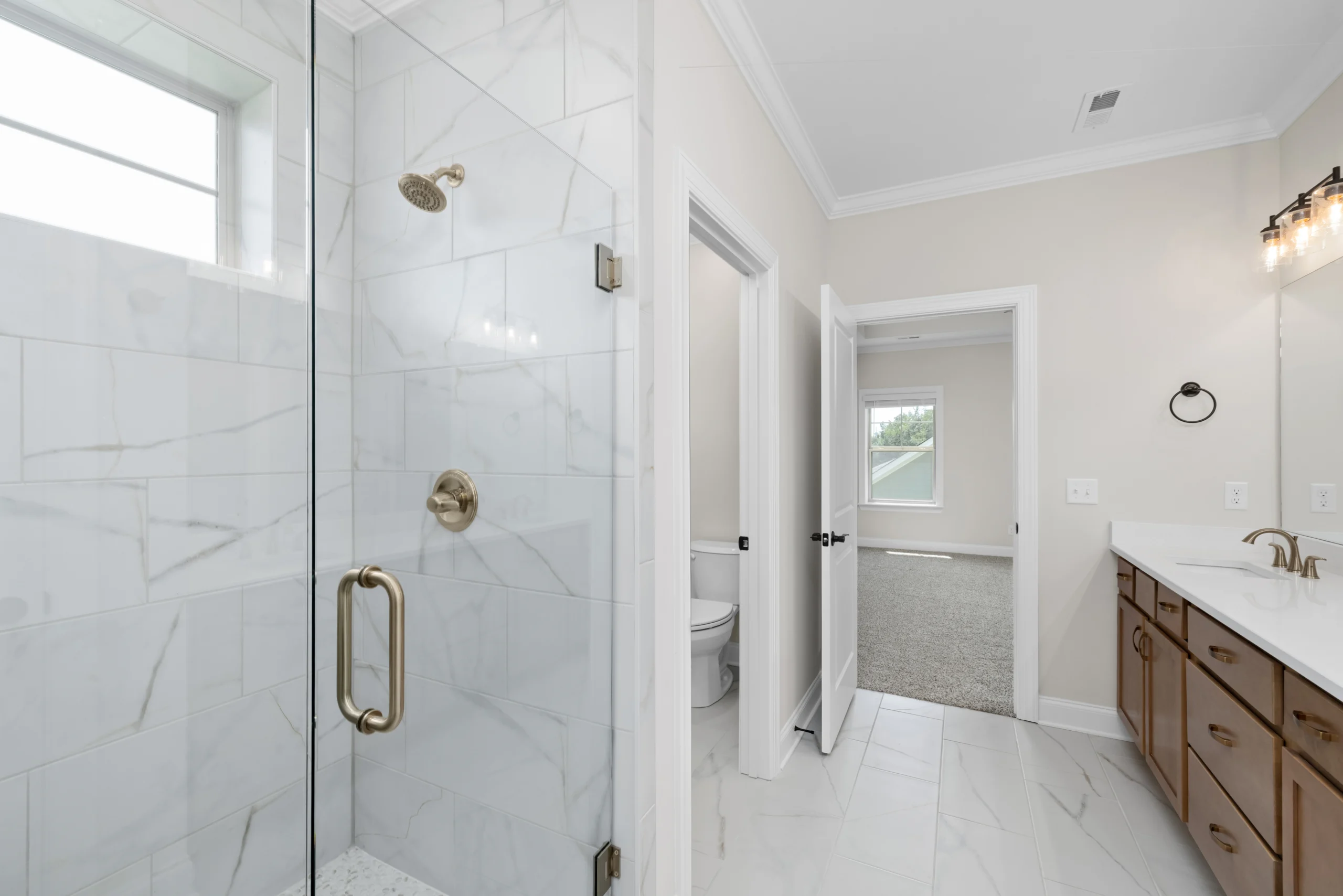 Bathroom with marble tiles, glass shower, wooden vanity, and white doors leading to a carpeted room. Pratt Home Builders, Chattanooga TN.