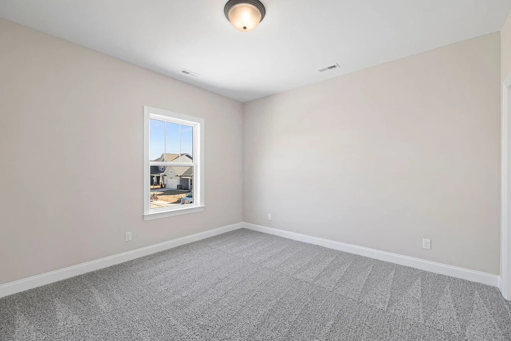 Empty room with light gray carpet, beige walls, and a ceiling light. A window reveals a suburban street view. Pratt Home Builders in Chattanooga TN.