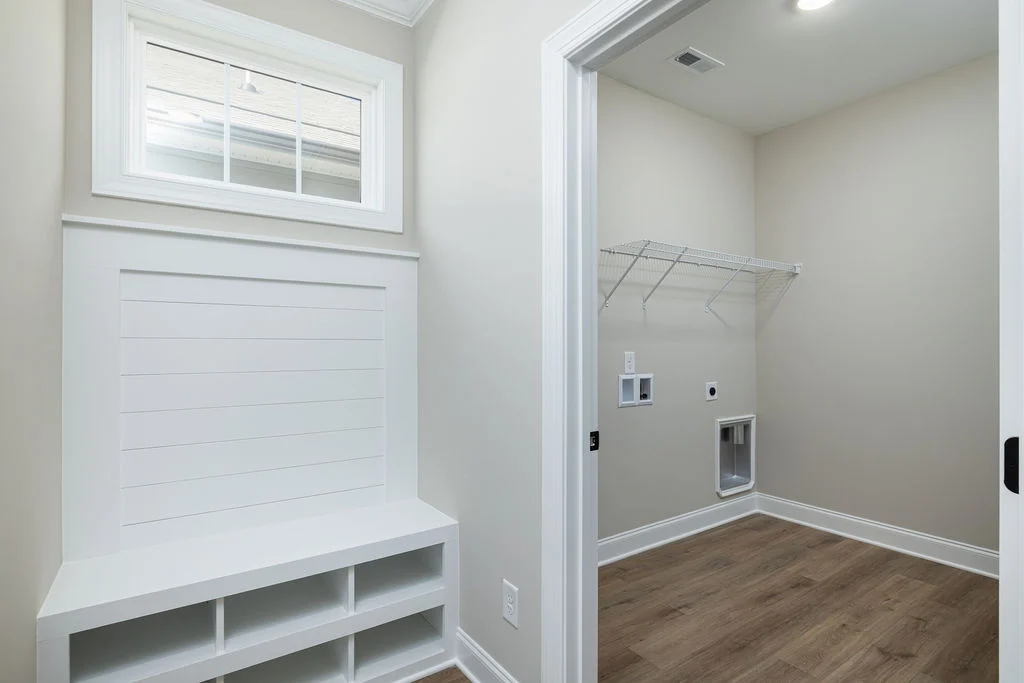 Mudroom with storage bench, white shelving, and laundry hook-ups. Designed by Pratt Home Builders in Chattanooga TN. Light wood floor, beige walls.