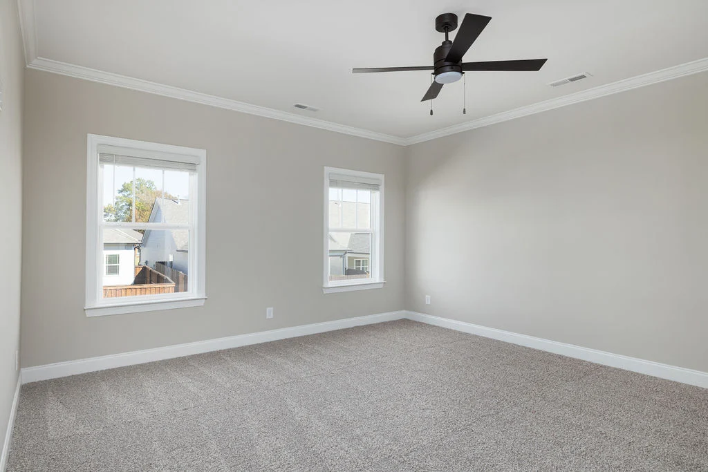 Empty room with beige walls, white trim, and carpet, featuring a modern ceiling fan and two windows. Built by Pratt Home Builders in Chattanooga TN.